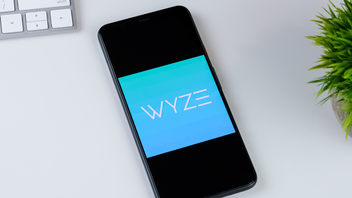 Logo de l'application Wyze sur un écran de smartphone, avec une plante et un clavier sur un bureau blanc en arrière-plan