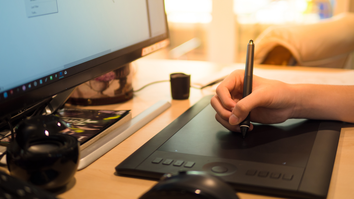Primo piano della mano di una persona che disegna su una tavoletta Wacom accanto al monitor di un computer