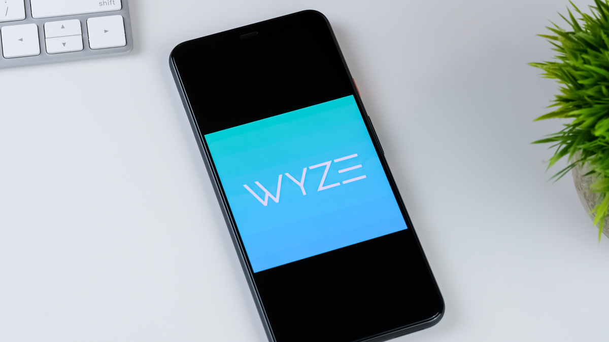 Wyze app-logo op een smartphonescherm, met een plant en toetsenbord op een wit bureau op de achtergrond