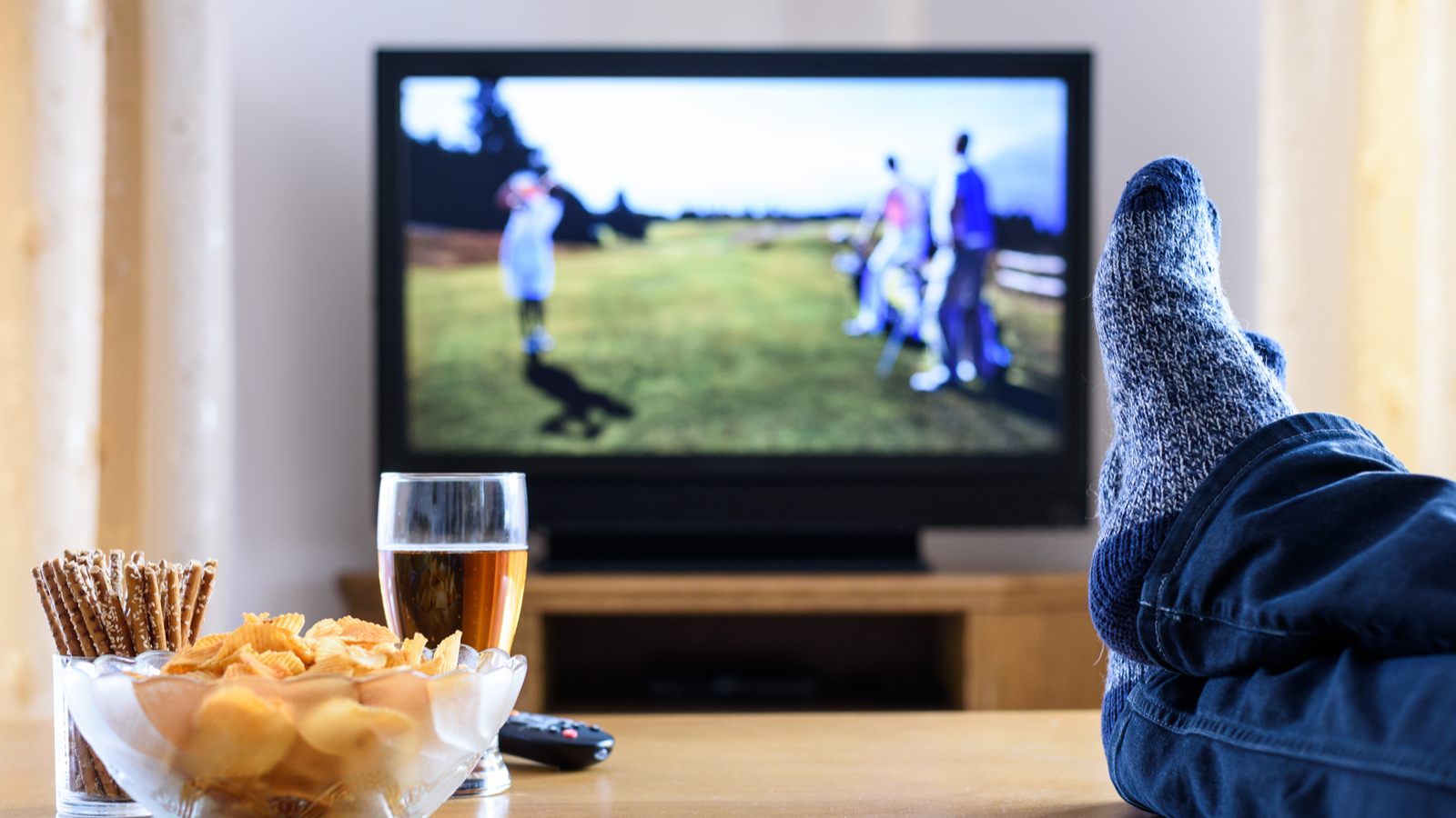 Televisie aan op de achtergrond, met gekruiste voeten op de salontafel naast snacks en bier op de voorgrond