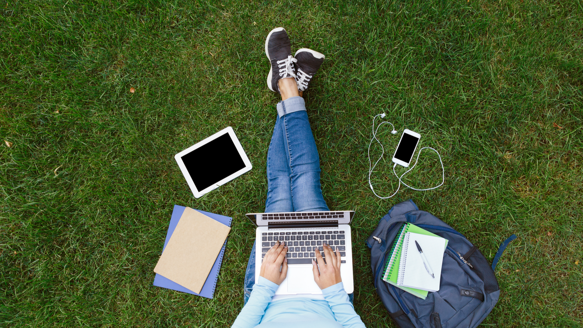 Bovenaanzicht van persoon zittend op gras in park met laptop, tablet, smartphone, hoofdtelefoons, notebooks en rugzak