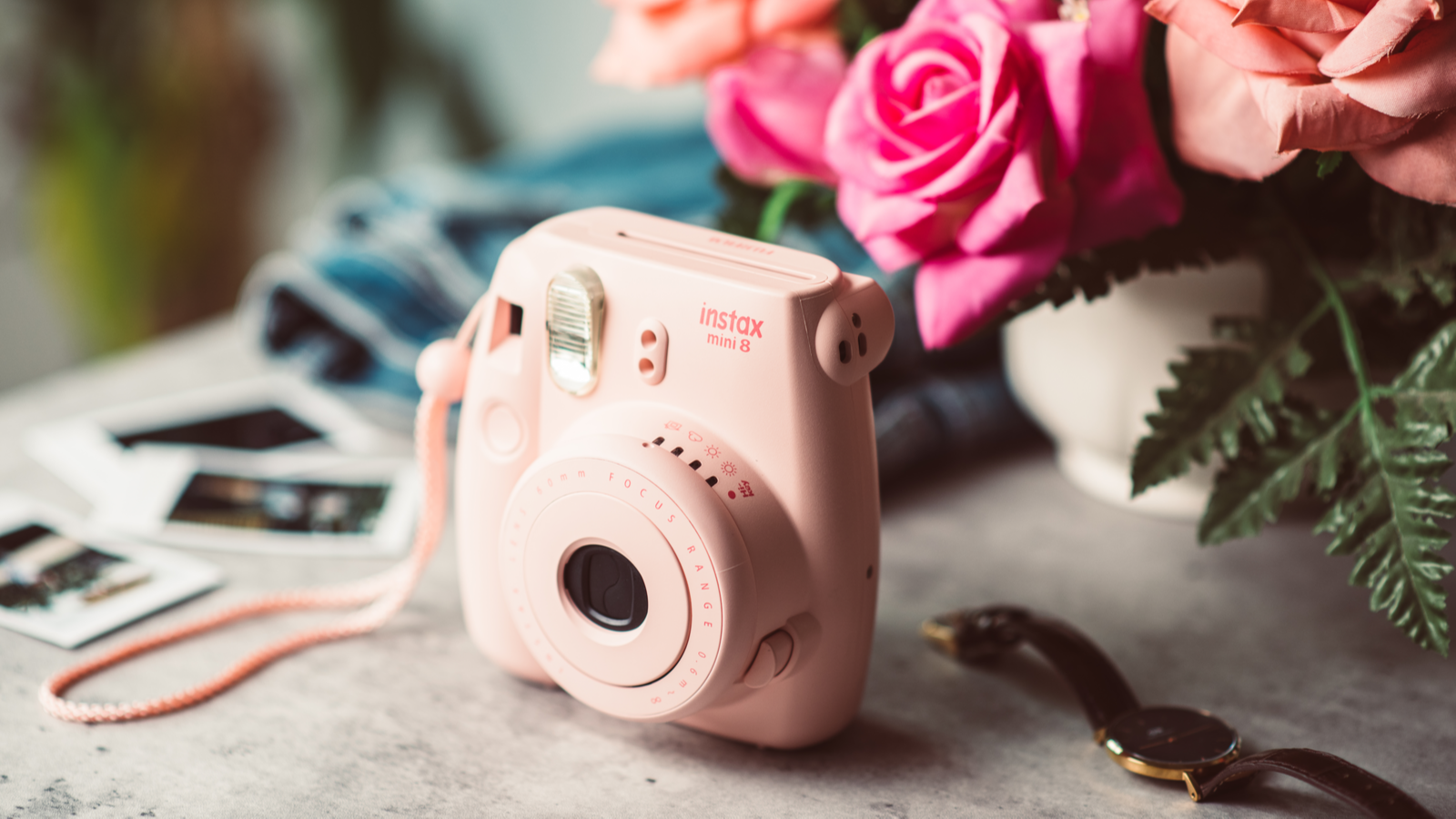 The pink Fujifilm Instax mini 8 on table next to photos, a watch, and a small vase of bright roses