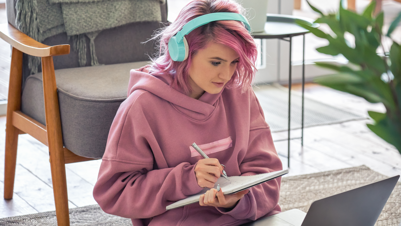 Người tuổi đại học với mái tóc hồng đeo tai nghe và ghi chú khi xem video trên máy tính xách tay
